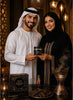 A man in a white kandura and sheath cap and a woman in a black abaya with gold embroidery hold a small black jar together. A branded product box reading 'Ole&Hana' and 'Al Mawlid' sits on a table with decorative lanterns and a incense burner in a warm, ornate interior.