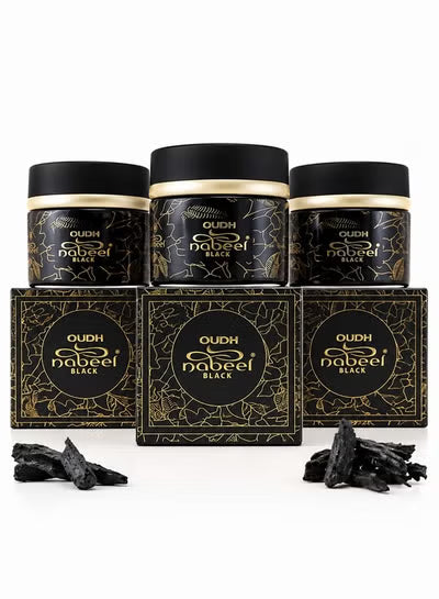 Three black cosmetic jars with gold detailing are arranged behind three matching rectangular boxes on a white background, with small pieces of black charcoal placed in front. The labels read 'OUDH nabeel BLACK'.